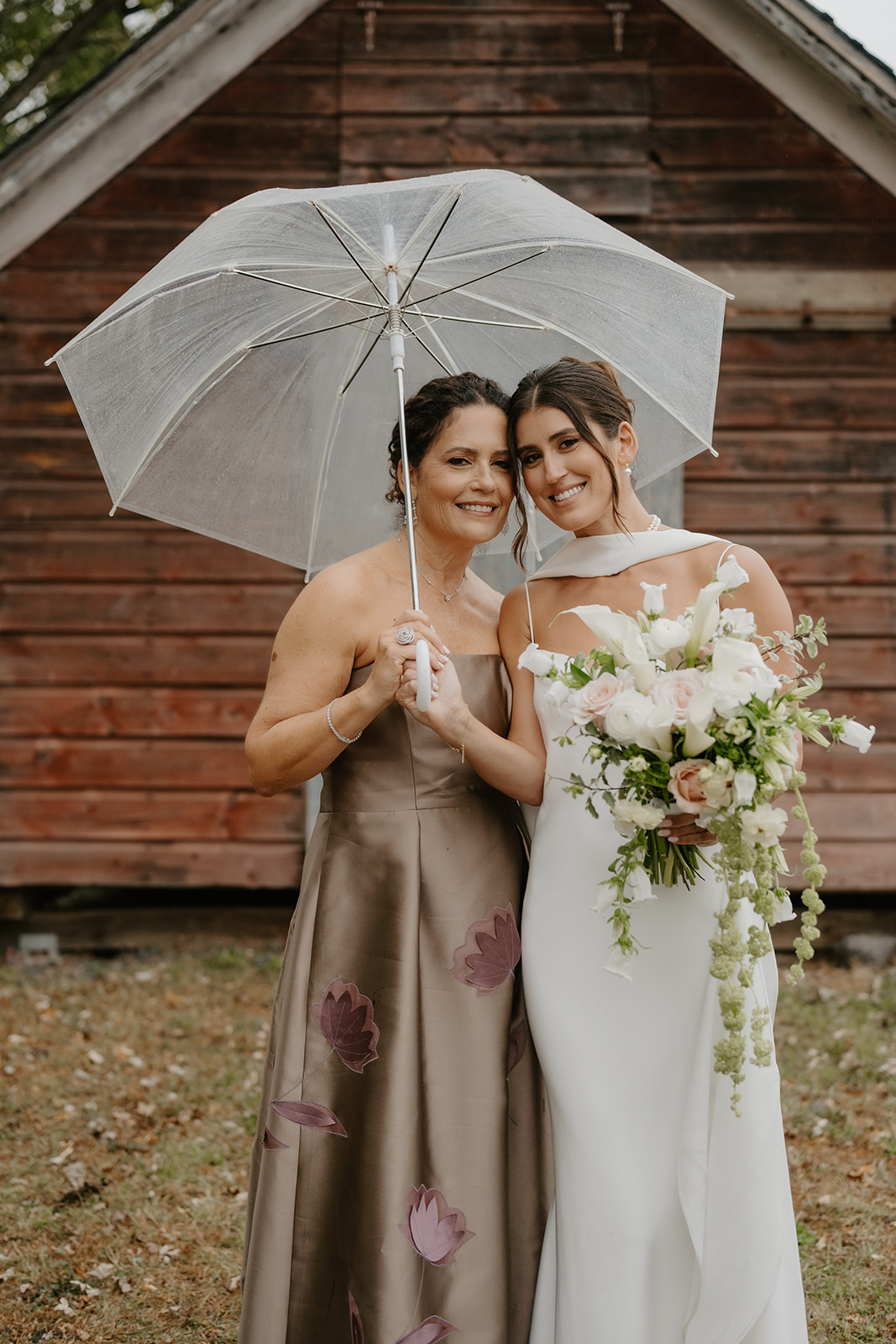 Bride and her mother smile under a clear umbrella in front of a rustic wooden shed.
