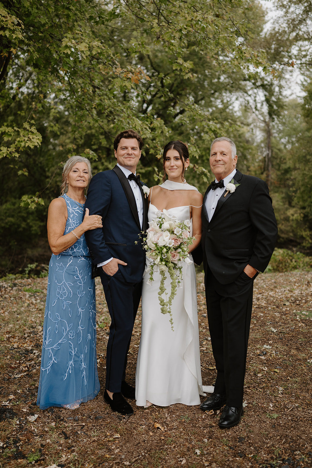 Bride and groom pose for a portrait with the groom's parents in a wooded setting.