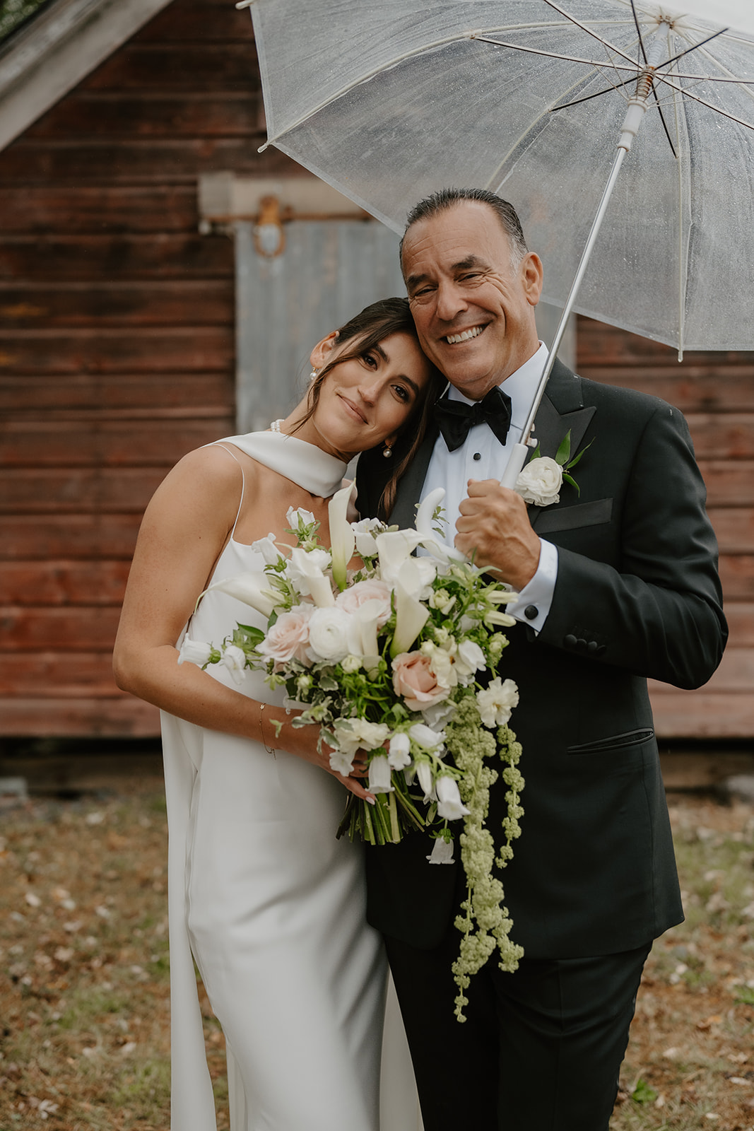 Bride leans her head on her father's shoulder under a clear umbrella, both smiling warmly.