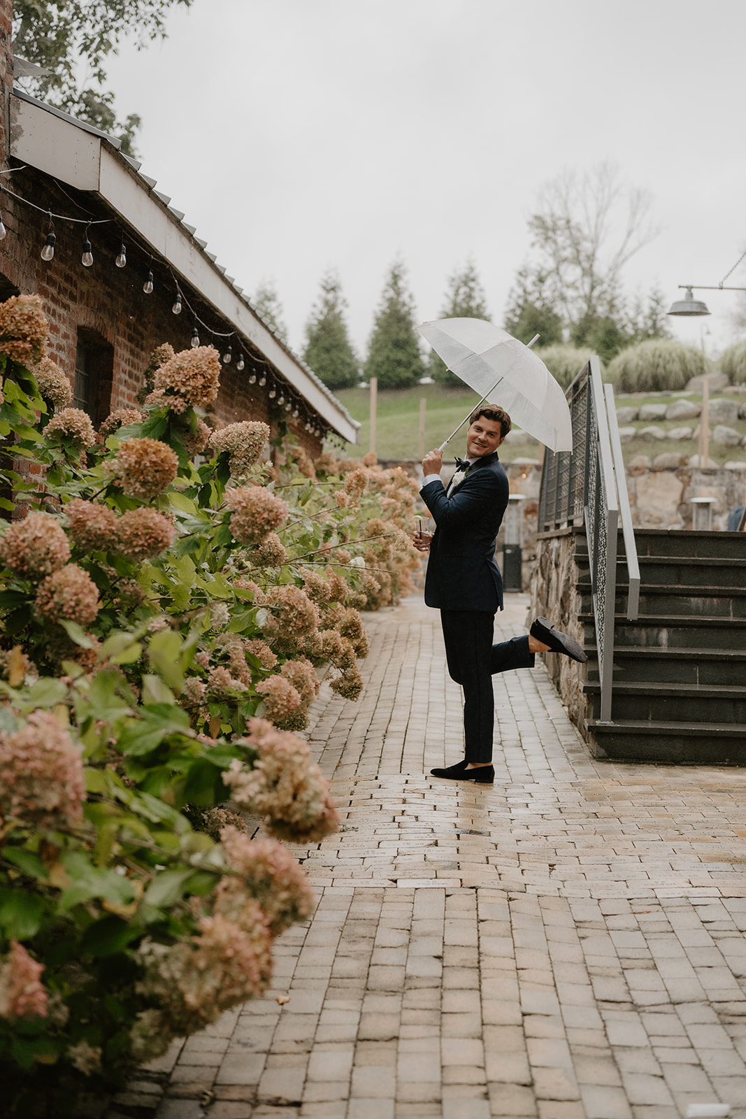 Groom poses playfully on one leg under a clear umbrella on a wet brick pathway by hydrangeas.