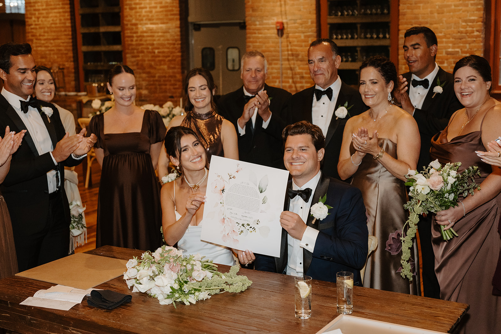 Bride and groom proudly present their signed Ketubah as family and friends applaud at the table.