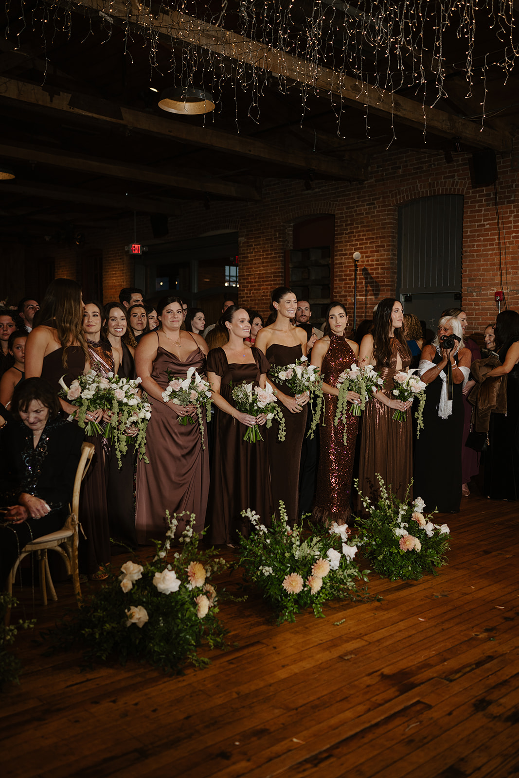 Bridesmaids in diverse brown and bronze gowns hold bouquets at the altar during the ceremony.