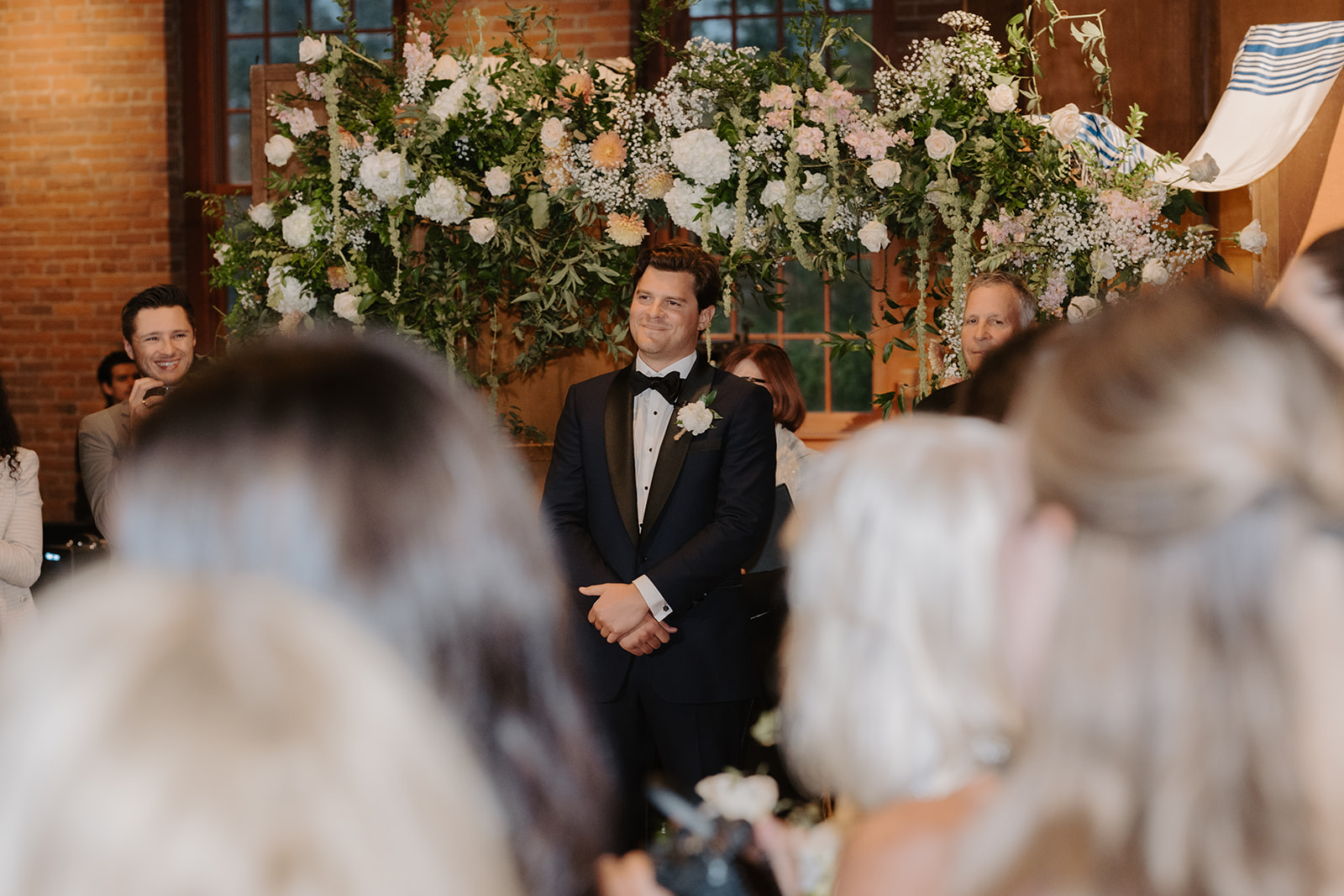 Groom smiles warmly from the altar under a lush floral Chuppah as guests look on.