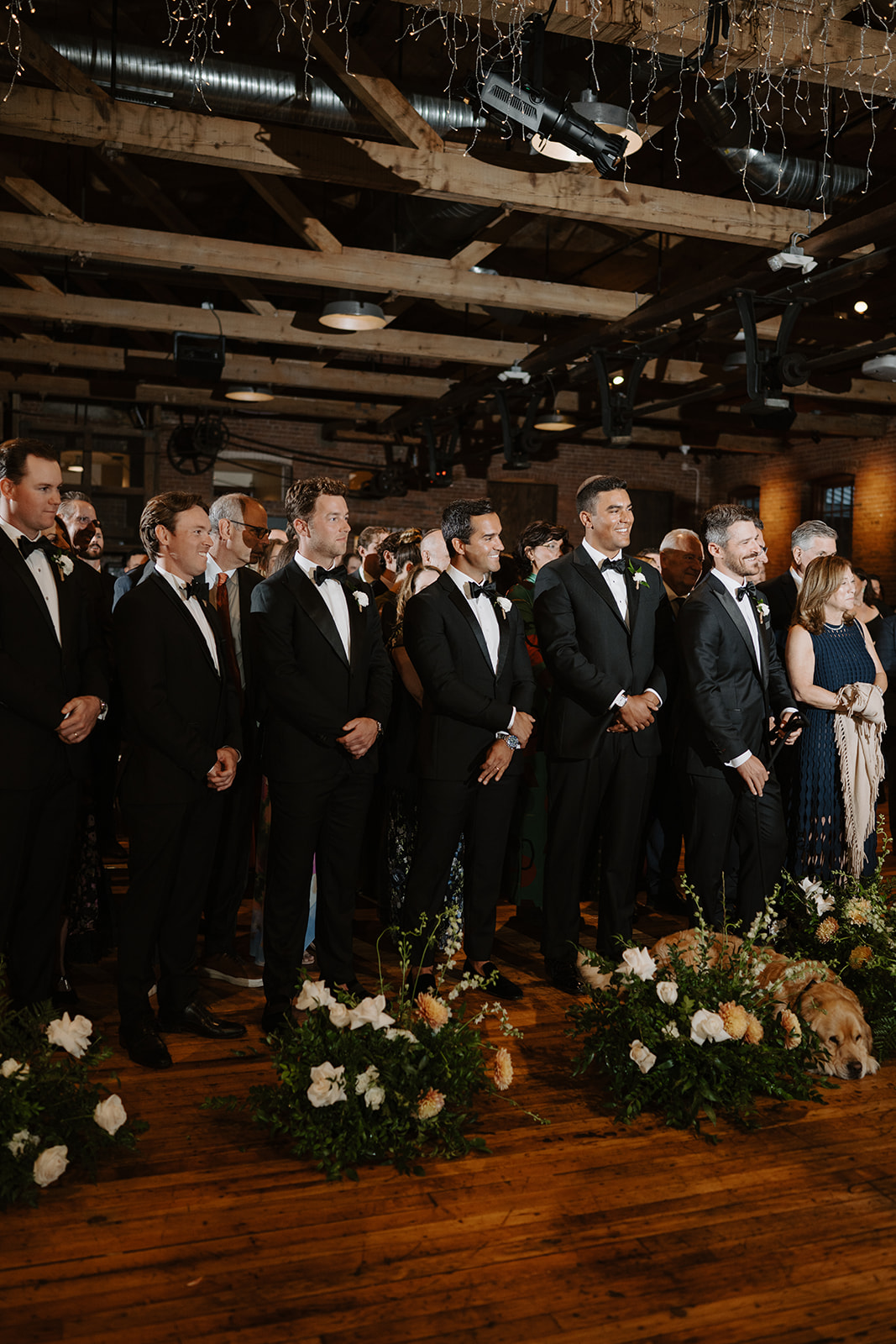 Groomsmen in black tuxedos smile during the ceremony as a golden retriever lies on the floor.