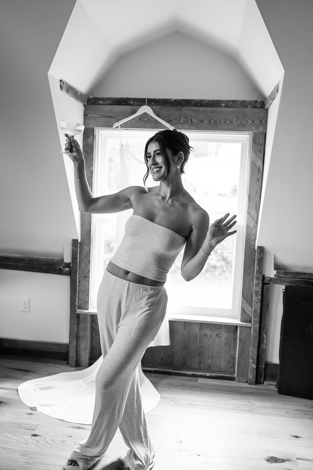 Black and white photo of the bride dancing playfully in loungewear with a champagne coupe.
