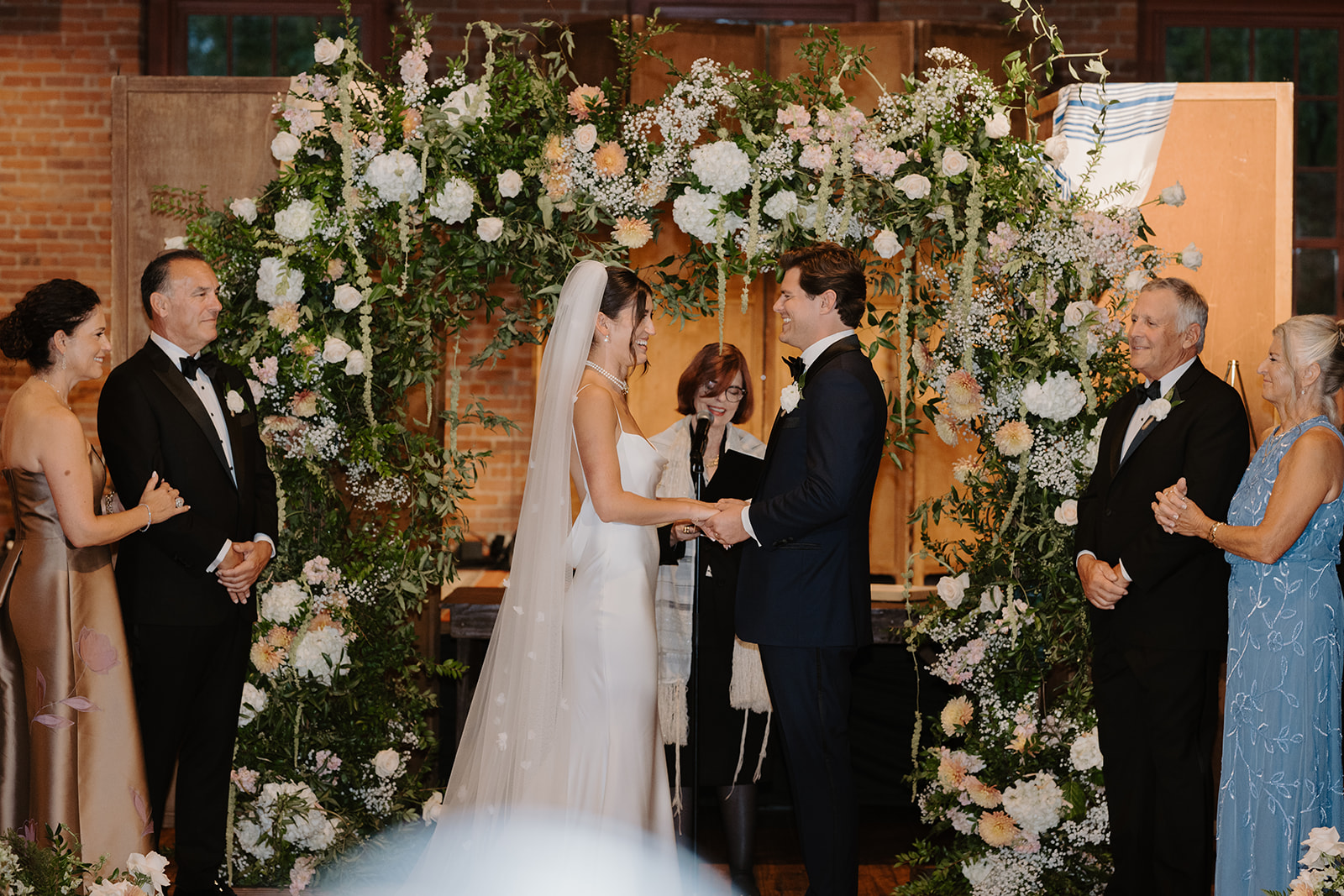 Bride and groom hold hands and smile during their ceremony under a beautiful floral Chuppah.