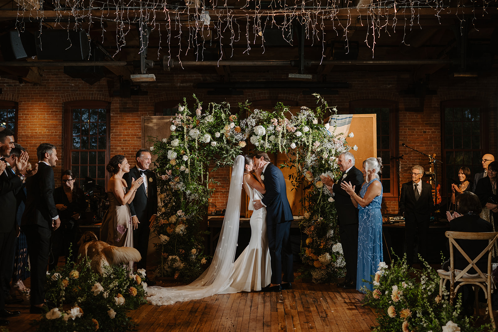 Bride and groom kiss during their wedding ceremony under a lush arch of baby's breath and greenery.