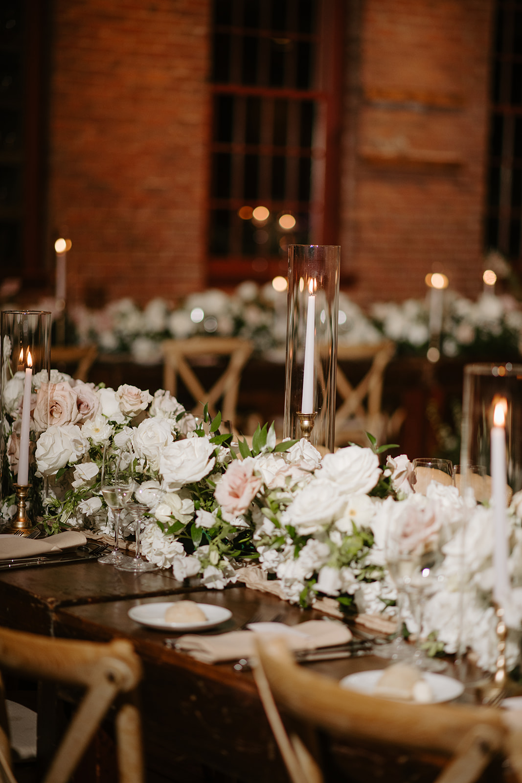 Lush white and blush floral runner with tall glass-encased taper candles on wooden tables.