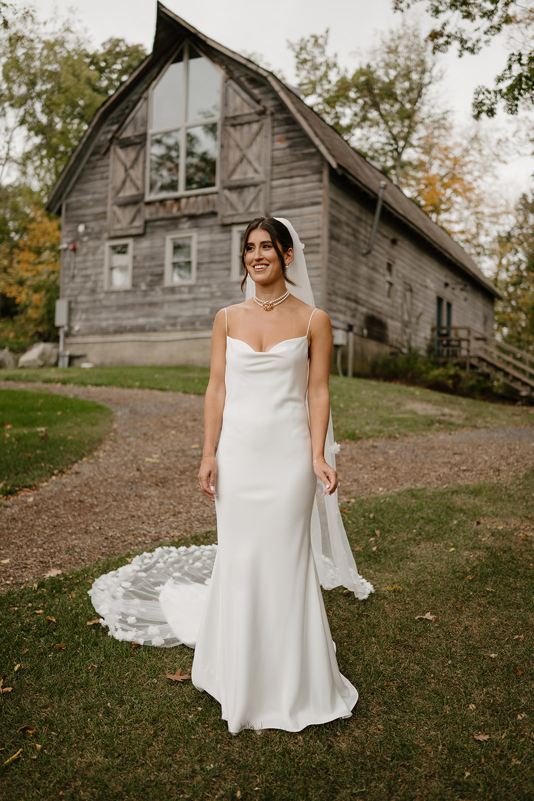 Bride in a white satin gown stands smiling on a grassy lawn in front of a rustic wooden barn.