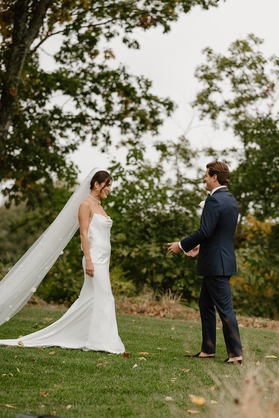 Groom in a blue tuxedo smiles warmly at his bride in a white gown during their first look on a grassy field.