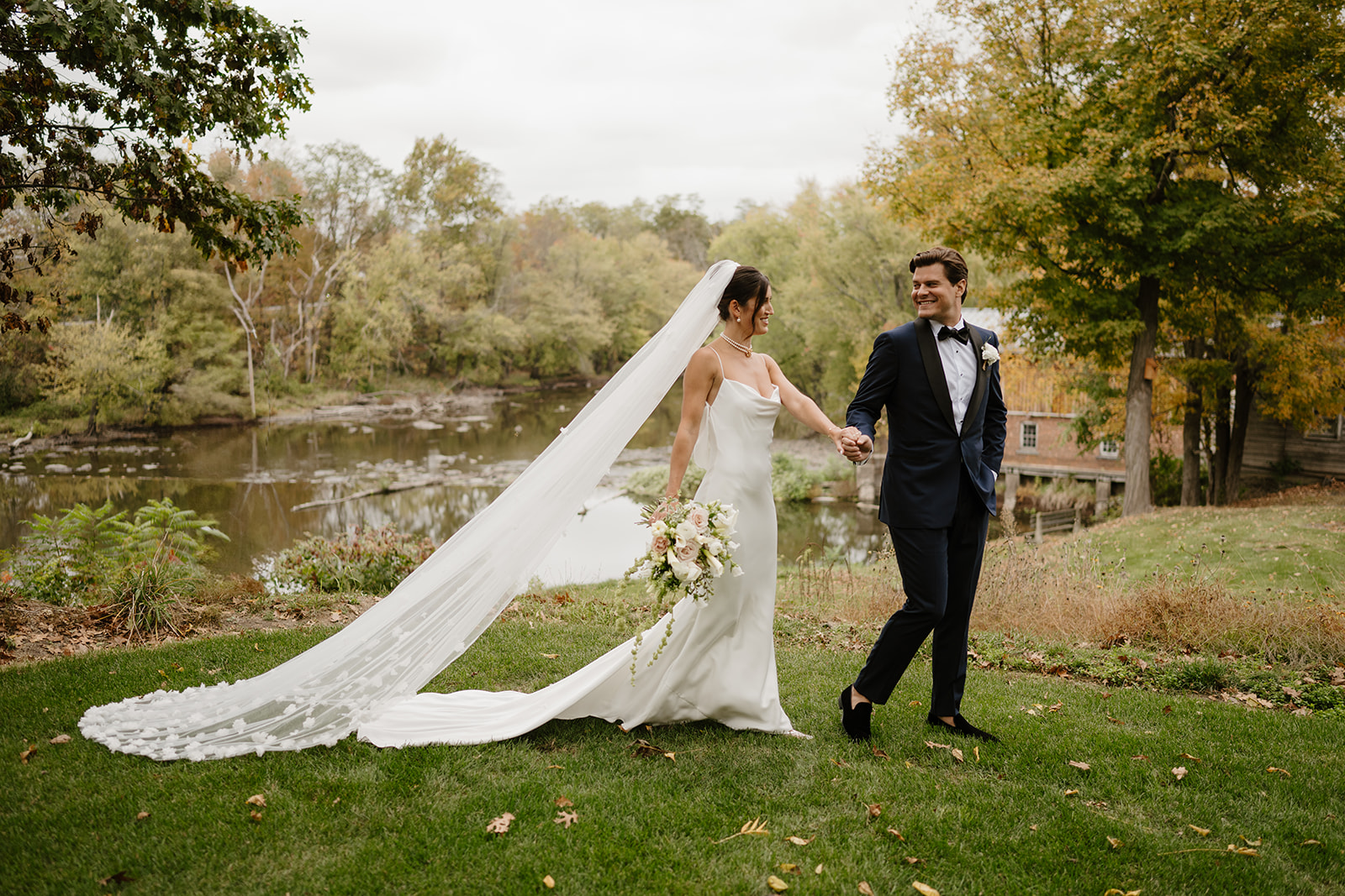 Bride and groom walk hand-in-hand smiling by a scenic river with autumn trees.