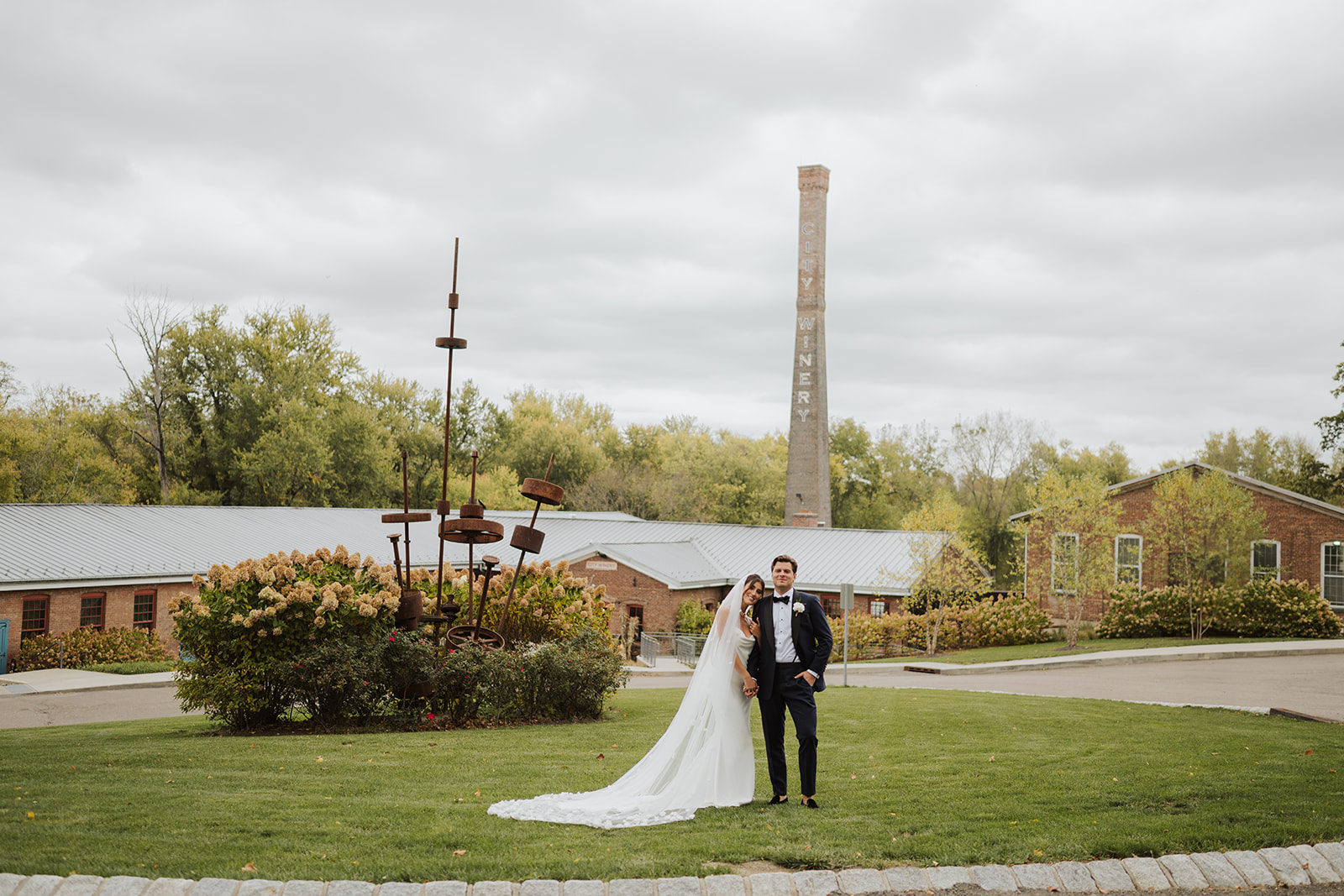 Bride and groom pose on the lawn at City Winery Hudson Valley with the iconic brick chimney behind them.