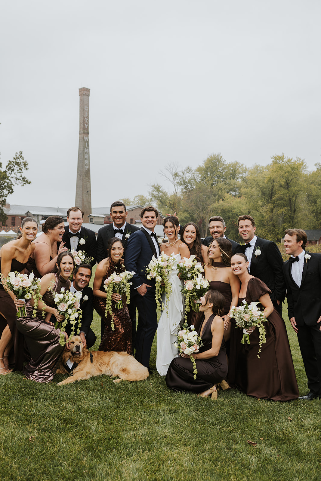 Wedding party and their golden retriever smile together on a lawn in front of a tall brick chimney.