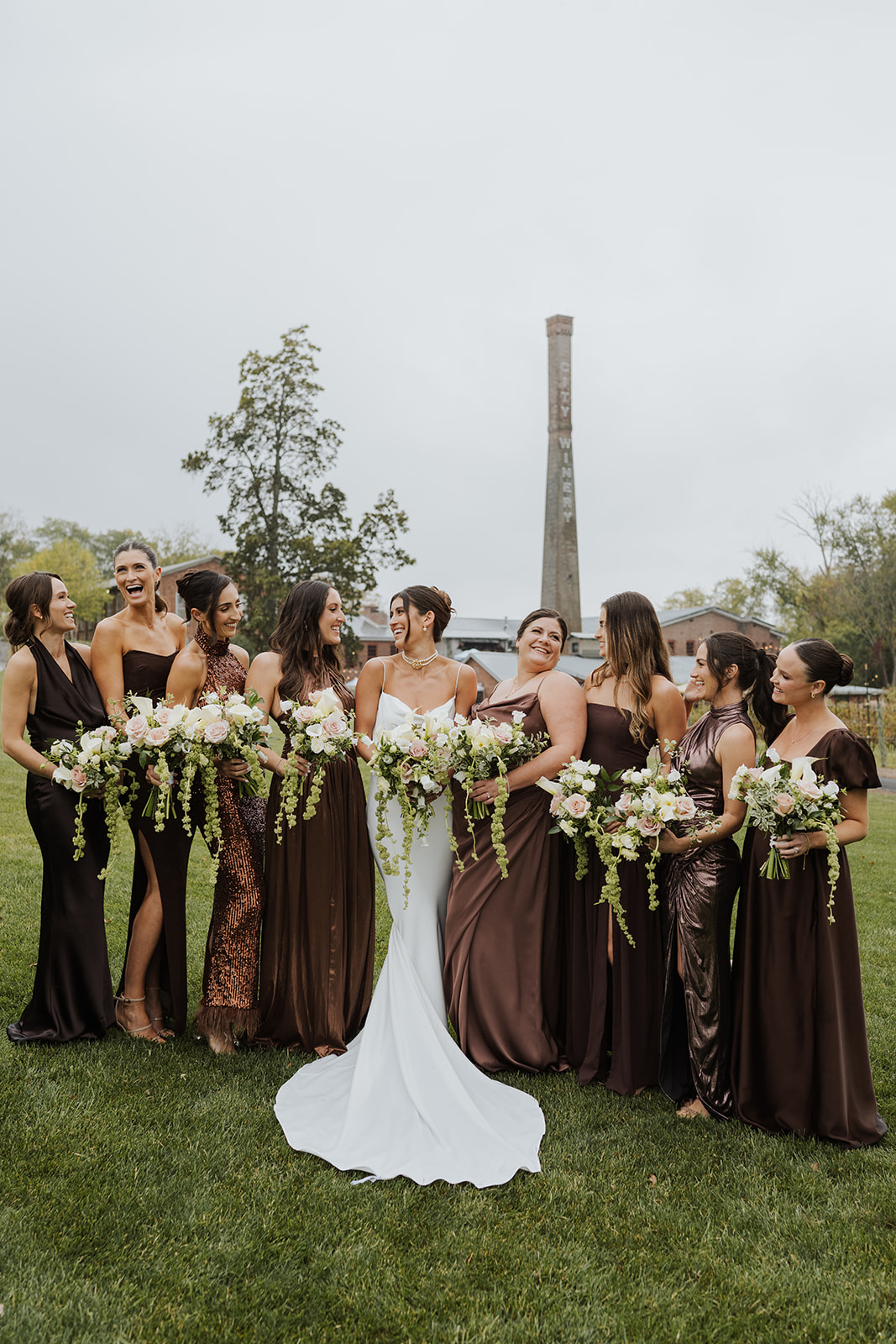Bride smiles at her bridesmaids dressed in shades of brown and bronze on a grassy lawn.