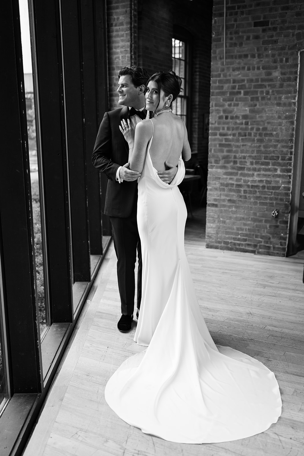 Black and white portrait of the bride looking back over her shoulder as the groom holds her by a window.