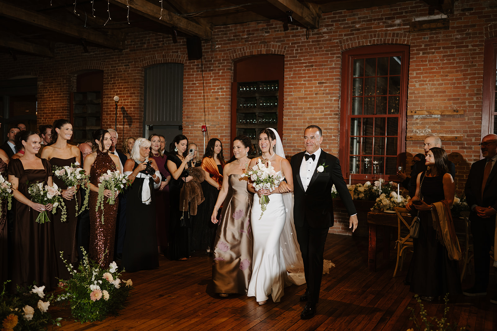 Bride walks down the aisle arm-in-arm with her mother and father inside a brick hall.