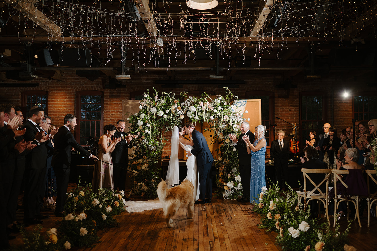 Bride and groom kiss under a floral arch during their ceremony as guests applaud and their dog watches.