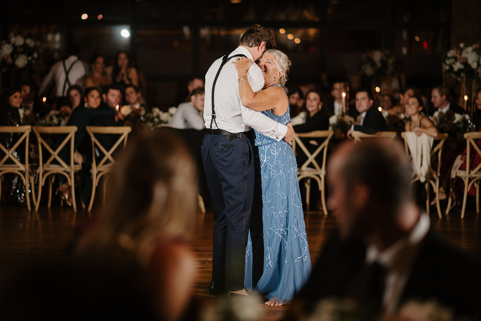 Groom hugs his mother warmly during their dance at the reception as guests look on.