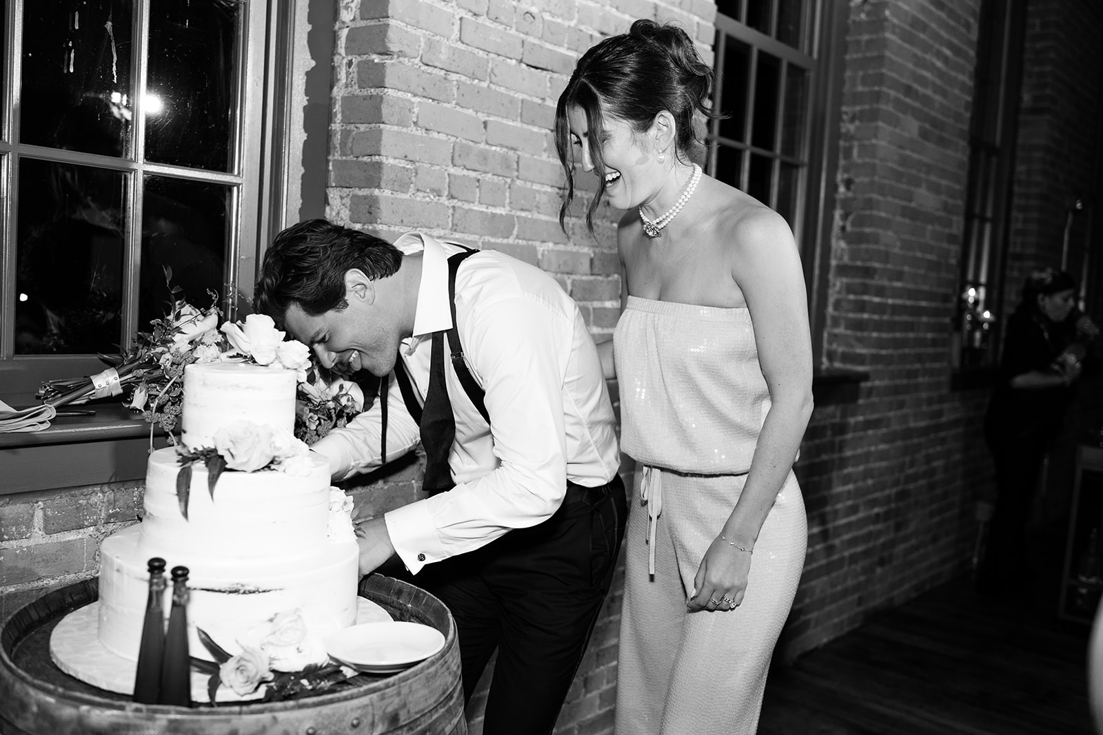 Black and white photo of the bride laughing as the groom playfully bites the wedding cake.