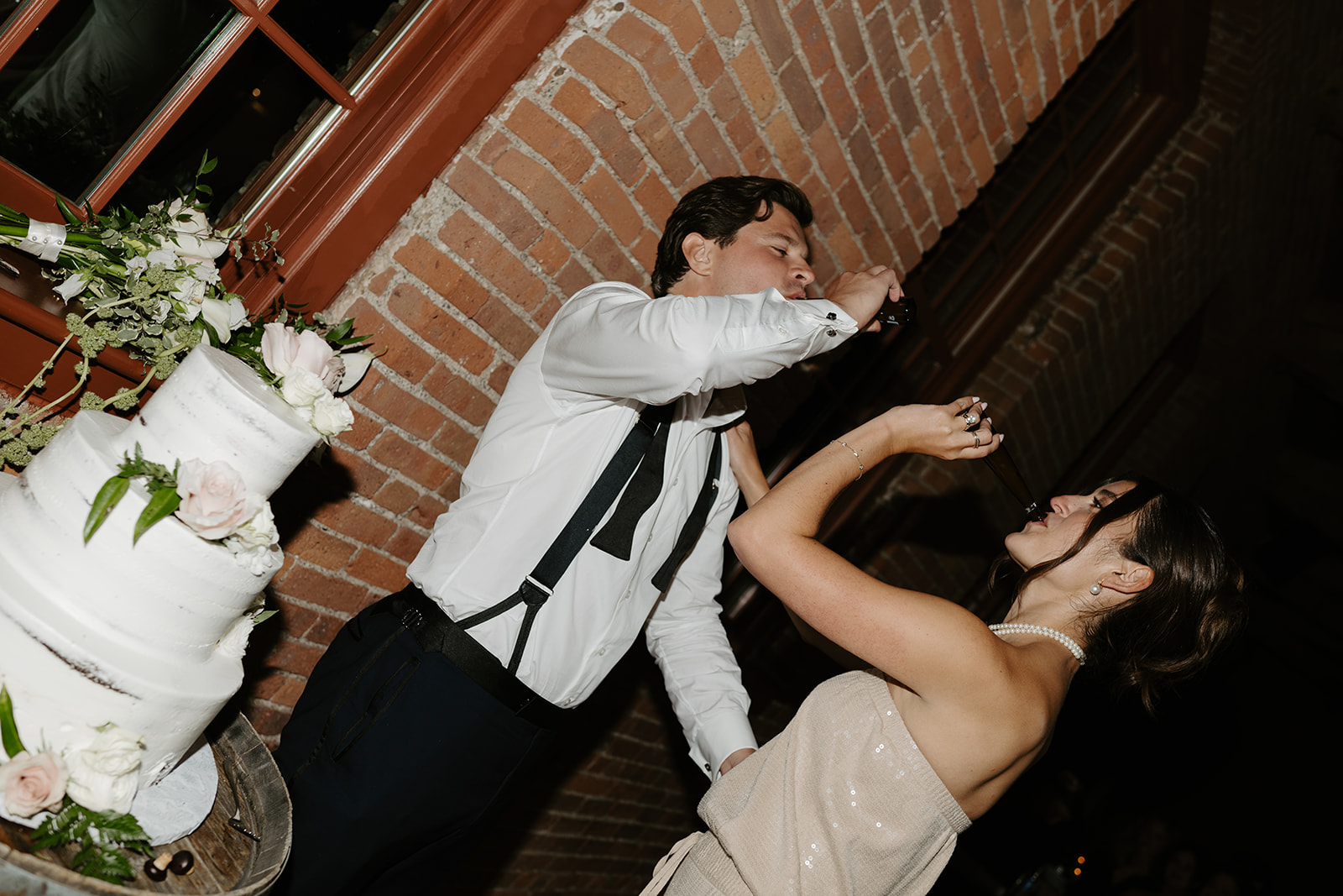 Bride and groom take celebratory shots from bottles next to their three-tiered wedding cake.
