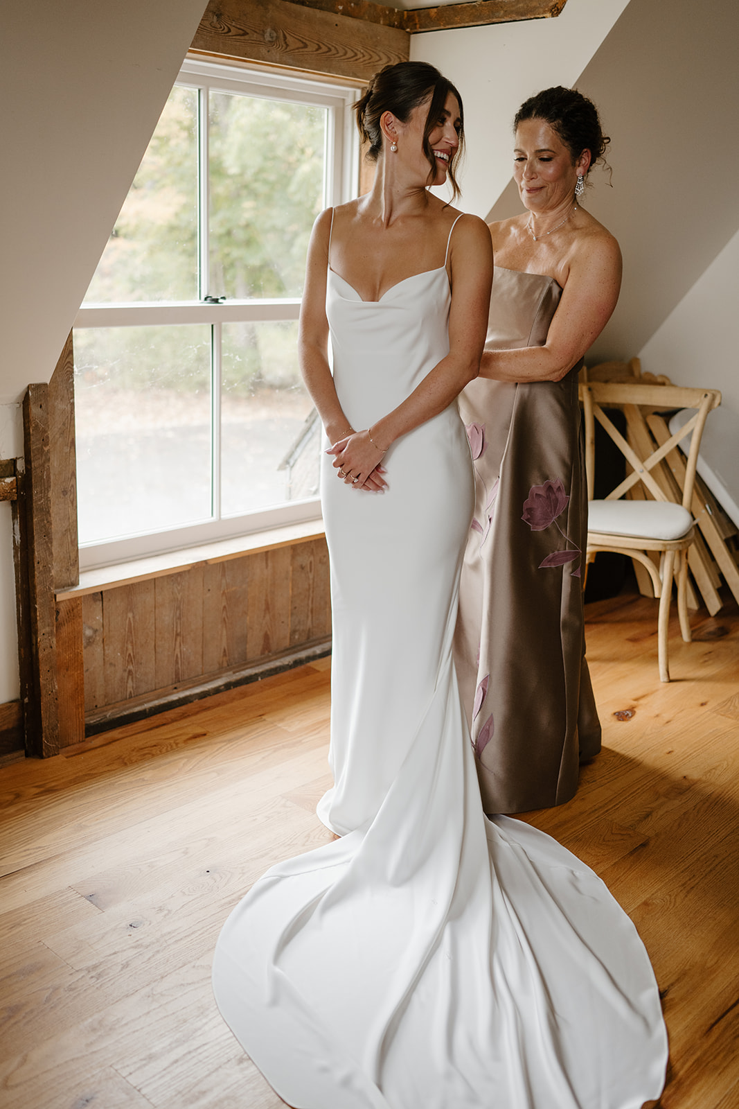 Mother of the bride helps her daughter dress in a sunlit loft with wooden floors.