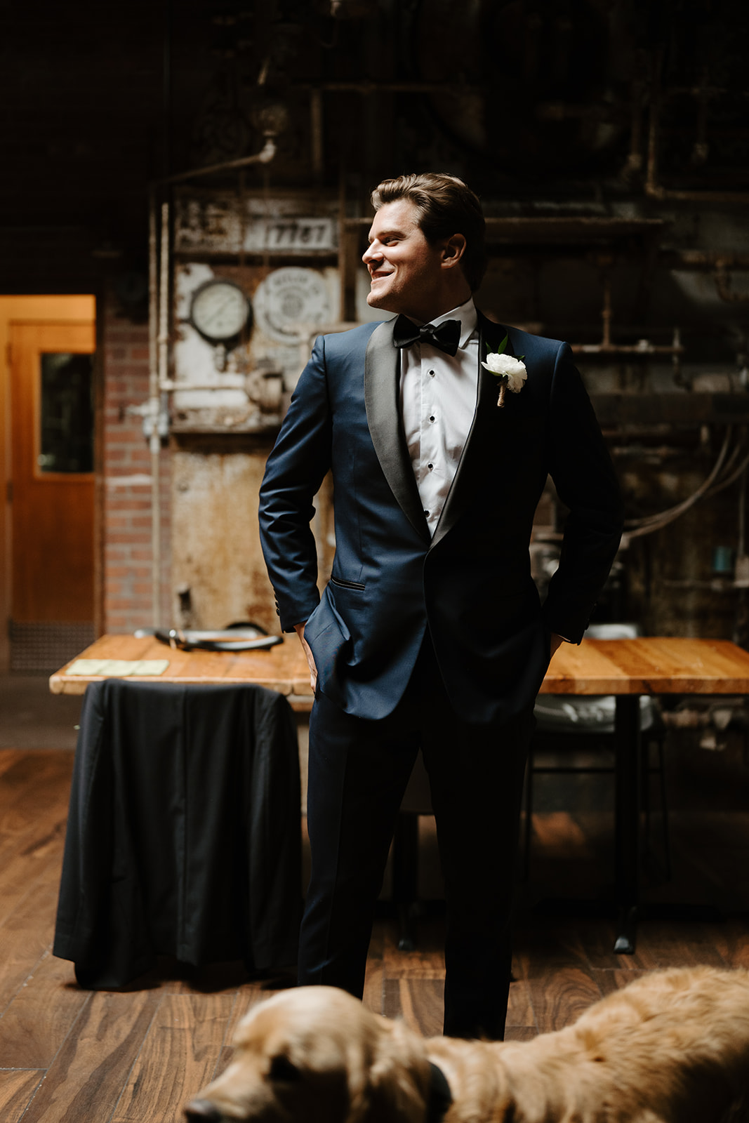 Groom in a blue tuxedo stands smiling in an industrial setting, with his dog in the foreground.