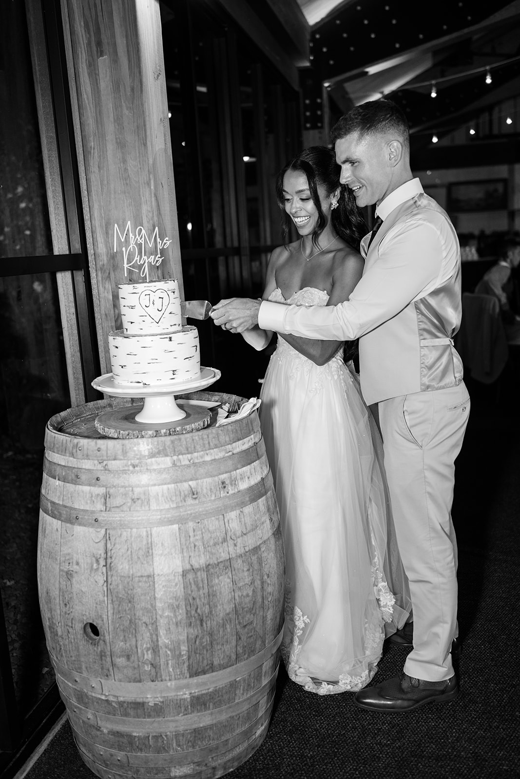 Black and white photo of the couple cutting their wedding cake on a wine barrel.