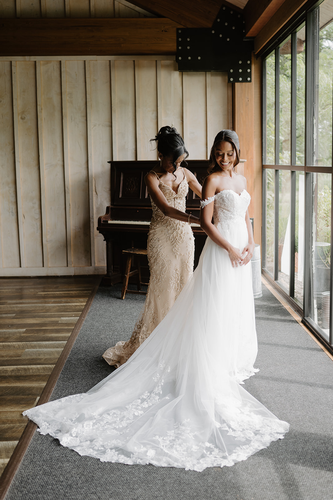 Bride and a bridesmaid stand together in the sunlit barn room, her lace train fanned across the floor.