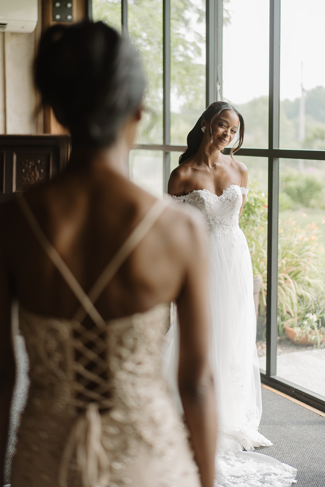 Bride in a corset-back gown smiles toward the light of a tall window.