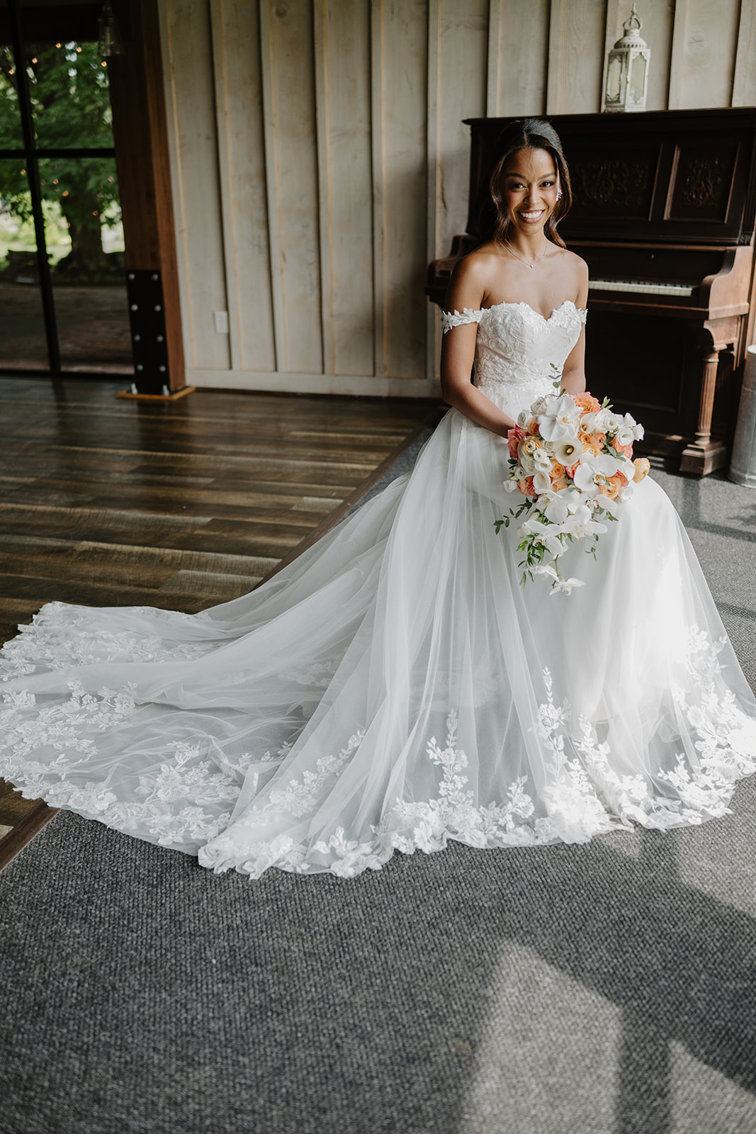 Bride poses beside an antique piano with her lace train spread across the wooden floor.