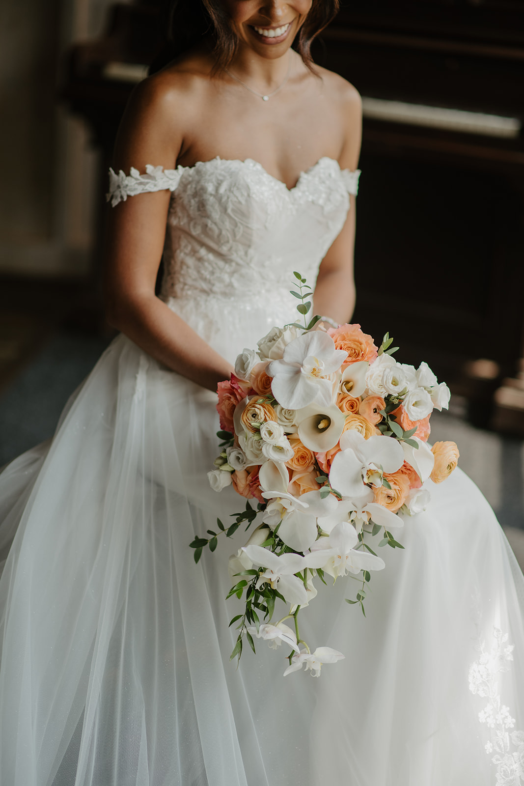 Bride in an off-the-shoulder tulle gown holds a bouquet of peach roses and white orchids.