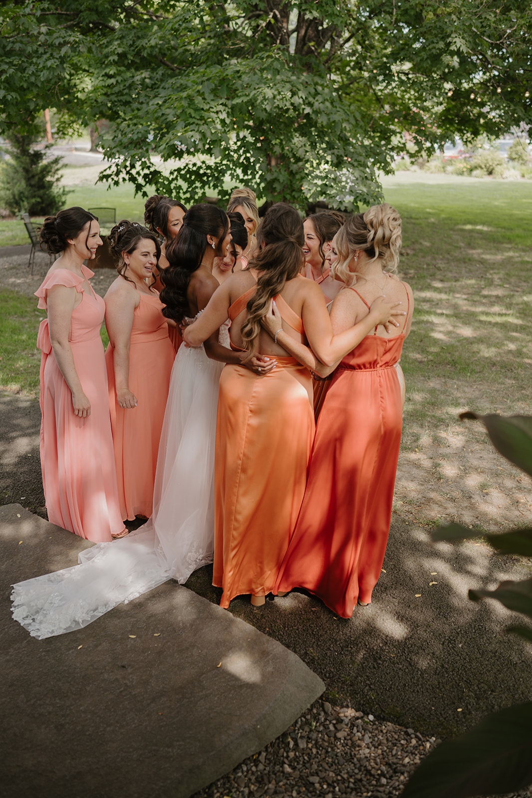 Bridesmaids in coral gowns circle the bride in a group hug beneath the trees.