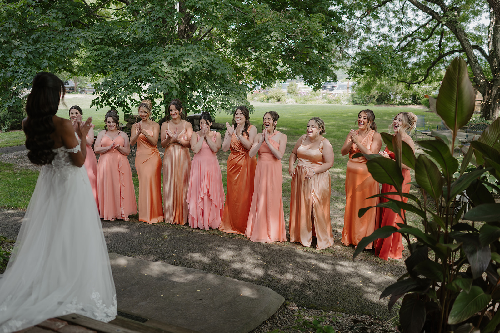 Bride approaches her bridesmaids in coral gowns waiting along the garden path.