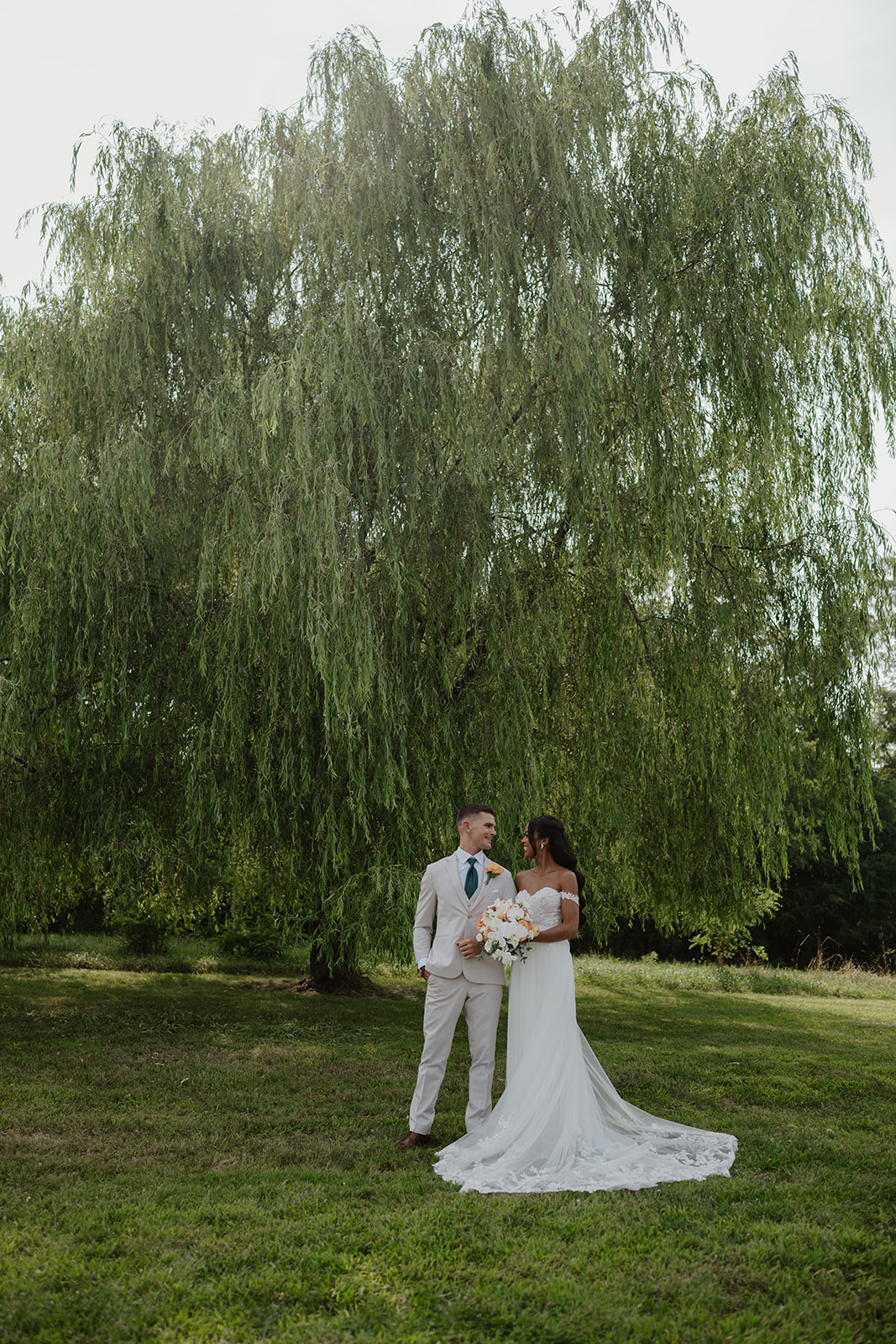 Bride and groom stand together beneath the wide branches of the willow tree.