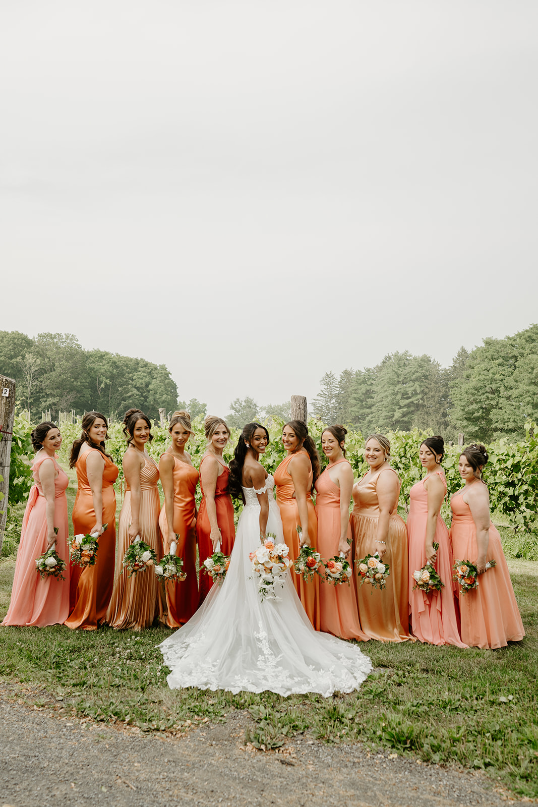 Bride stands at the centre of her bridesmaids in coral gowns before the vineyard.