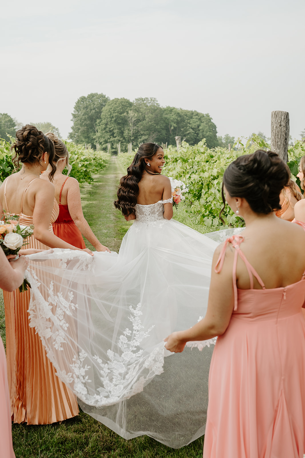 Bridesmaids carry the bride's lace train as she walks the vineyard path.