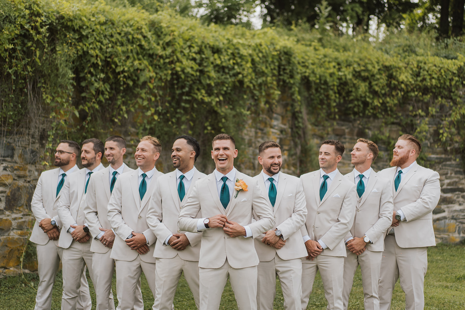 Groomsmen in cream suits and teal ties gather before the ivy-covered stone wall.