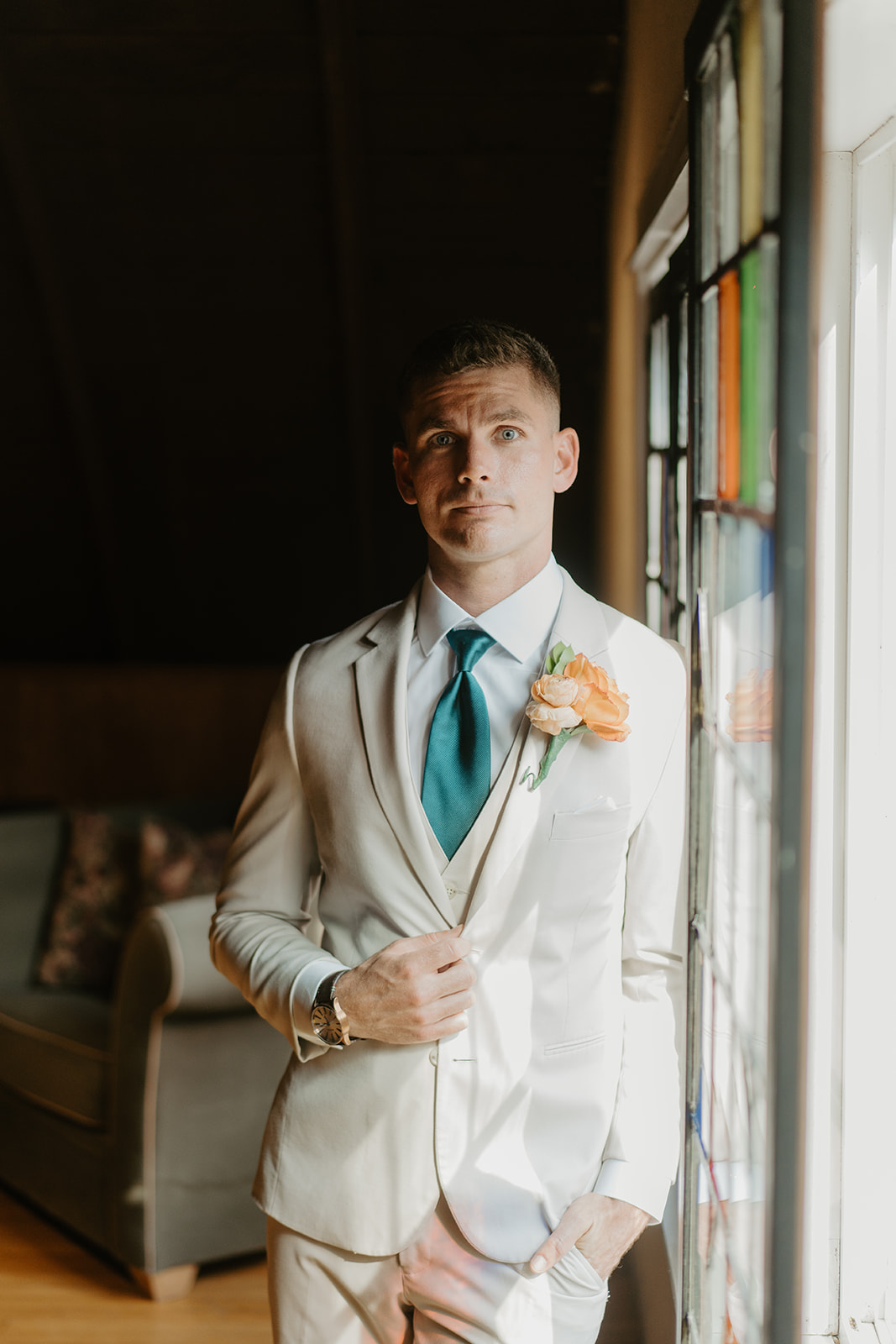 Groom in a cream suit and peach boutonniere waits beside the barn windows.