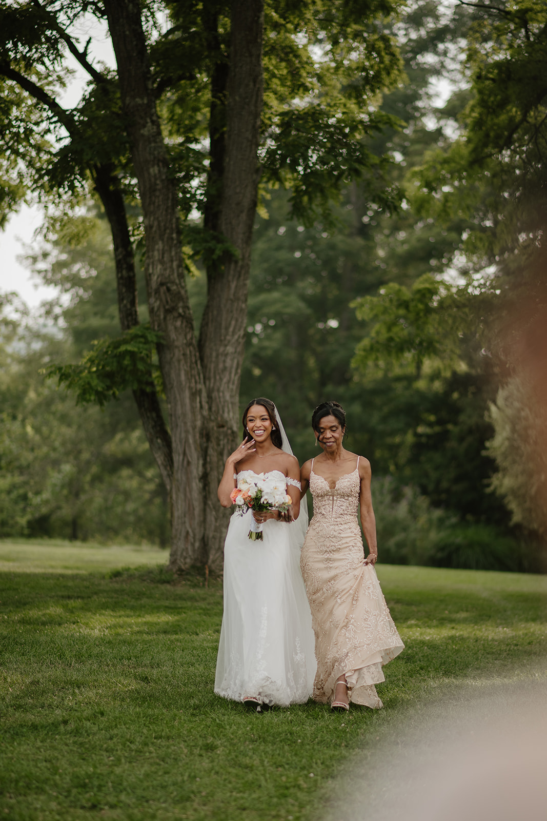 Bride walks the lawn with a bridesmaid, both carrying peach and white bouquets.