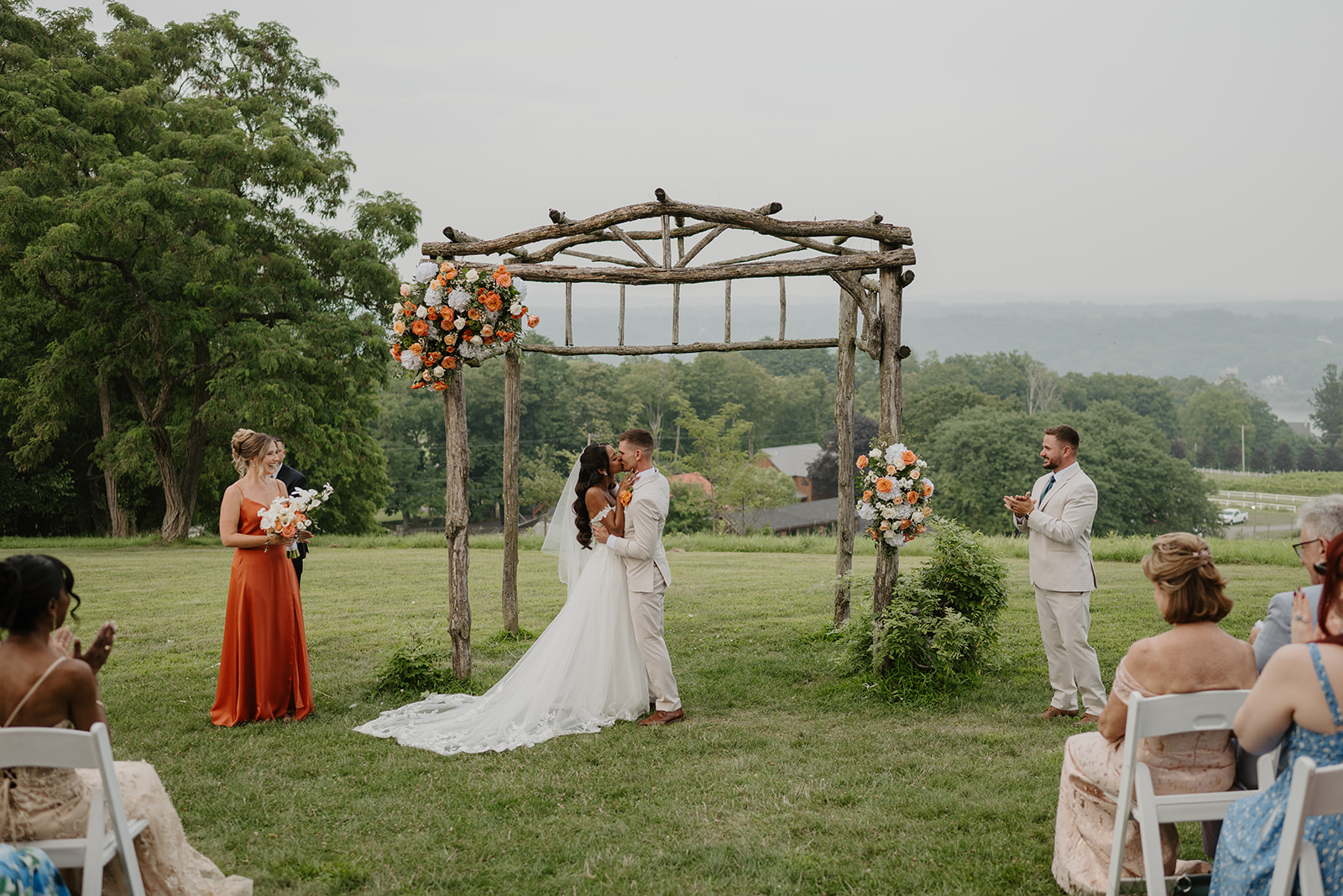 Bride stands at the floral arch during the hilltop ceremony as guests look on.