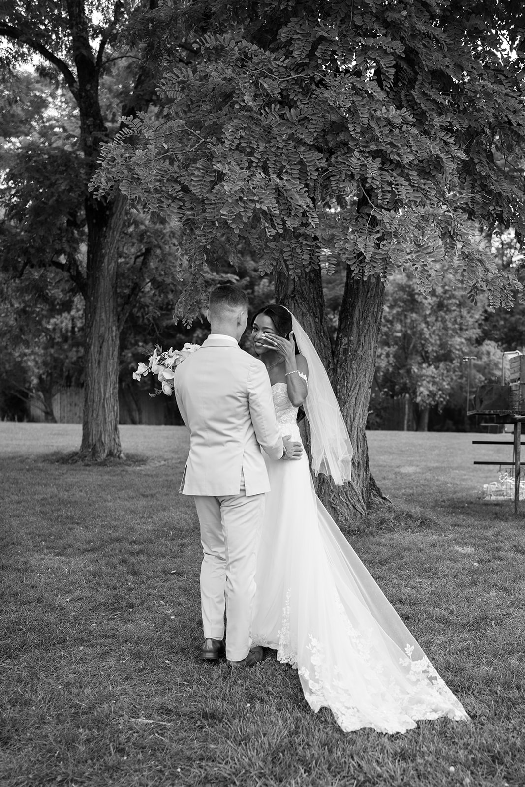 Black and white photo of the couple sharing a kiss beneath the trees, her veil trailing.