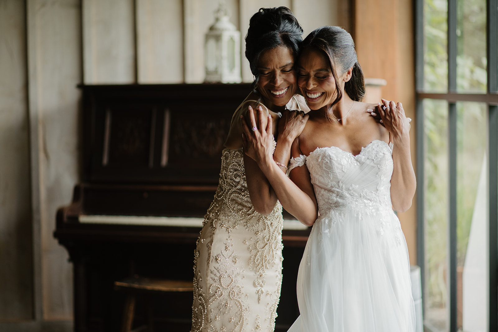 Bride and a bridesmaid in a gold beaded gown share a smile beside the antique piano.