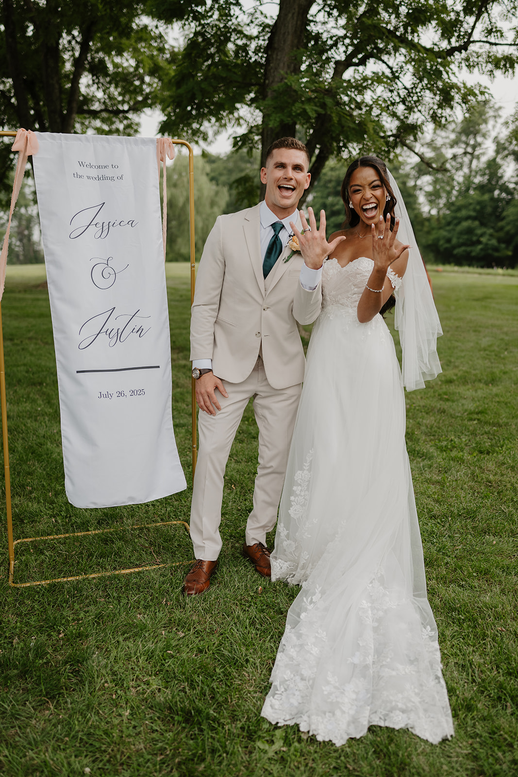 Newlyweds hold up their ringed hands beside their calligraphed welcome sign.