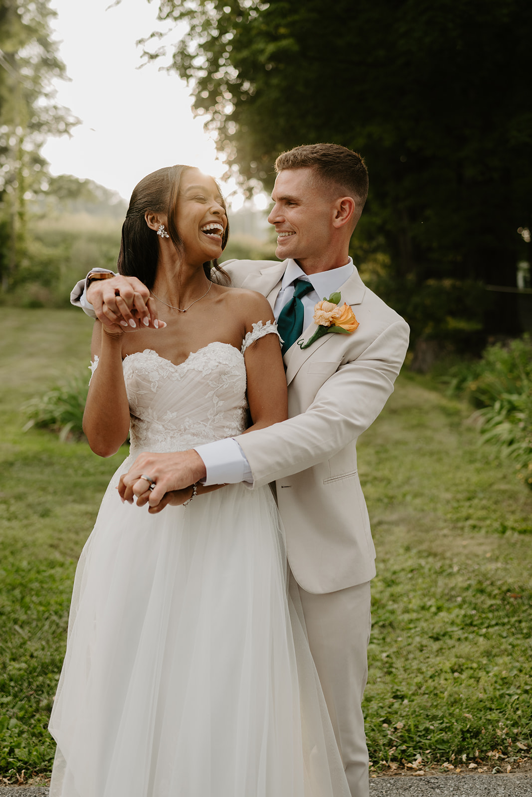 Groom wraps his arms around the laughing bride on the sunlit lawn.