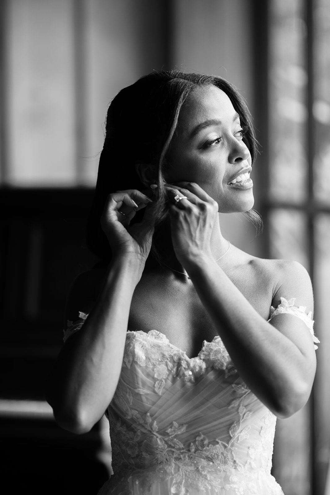 Black and white portrait of the bride adjusting her earring, framed by tall barn windows.