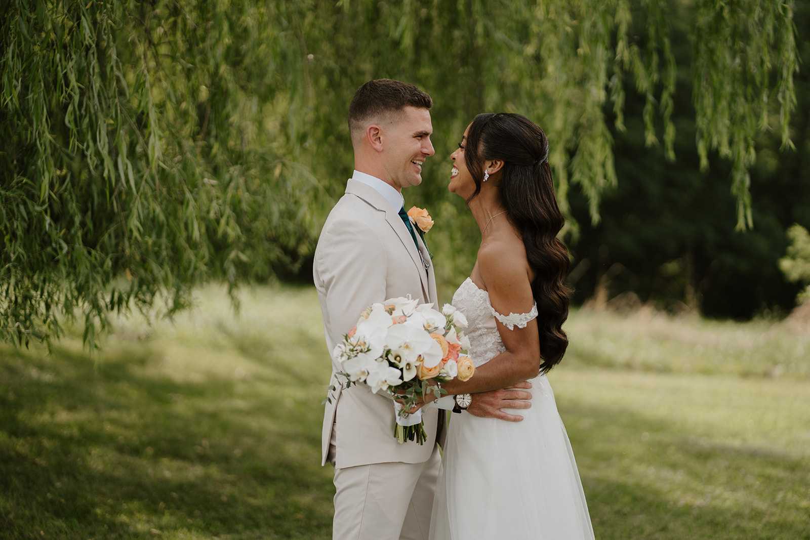 Bride and groom embrace under the willow tree, her bouquet at her side.