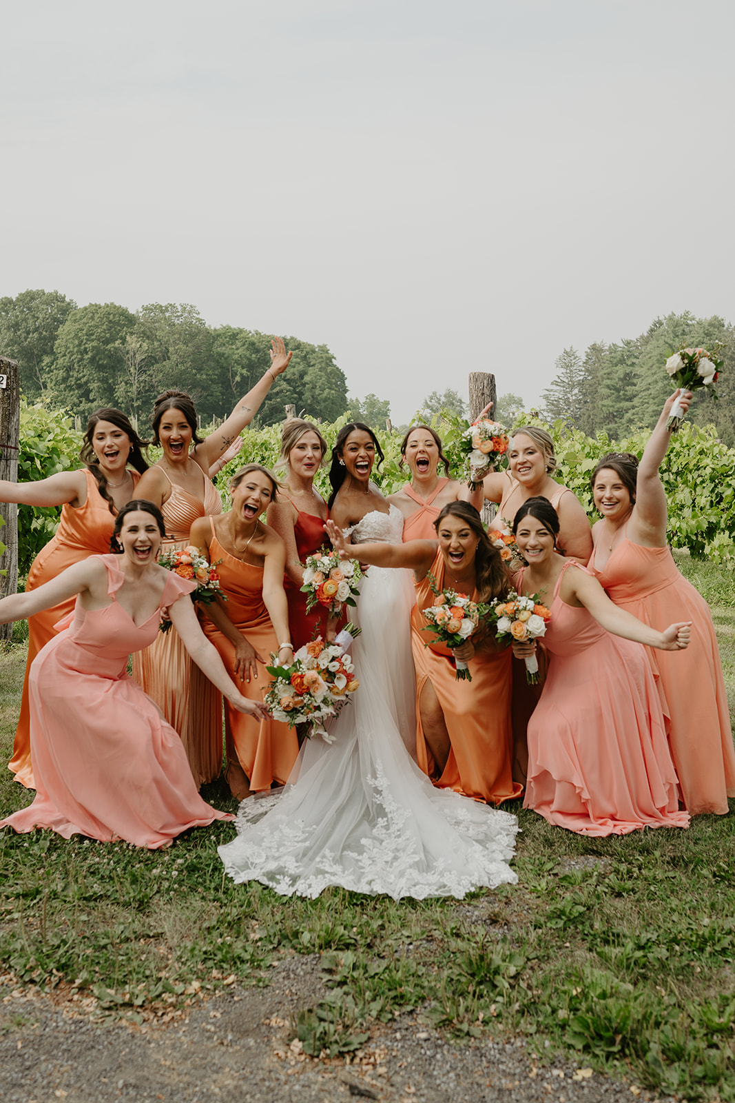Bridal party cheers around the bride in the vineyard, bouquets raised high.