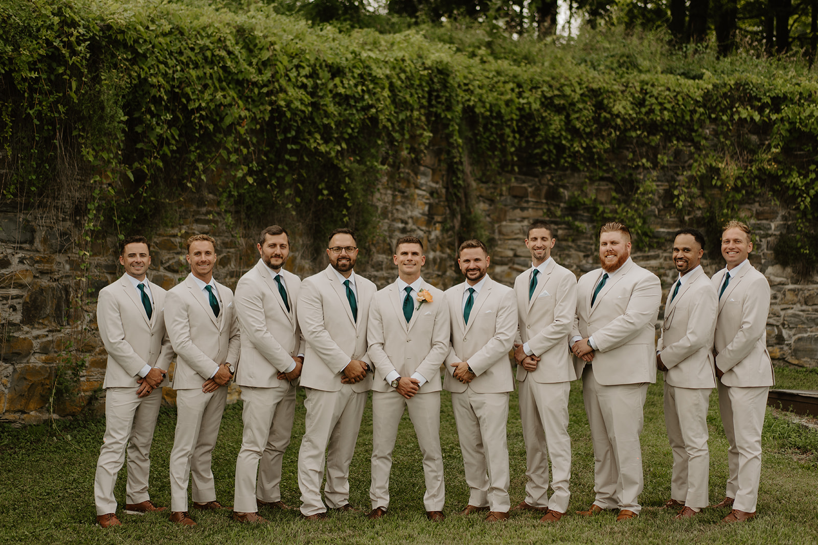 Groom and his groomsmen stand in a row before the ivy-covered stone wall.