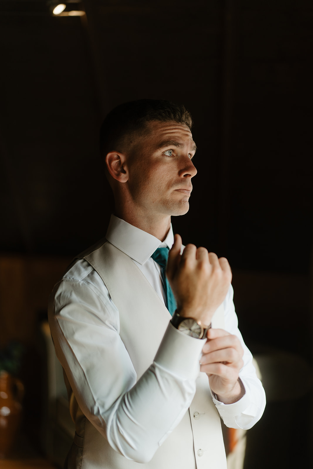 Groom adjusts his teal tie in warm window light before the ceremony.