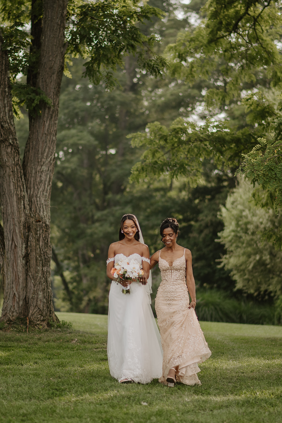 Bride walks the lawn with a bridesmaid, both holding peach bouquets.
